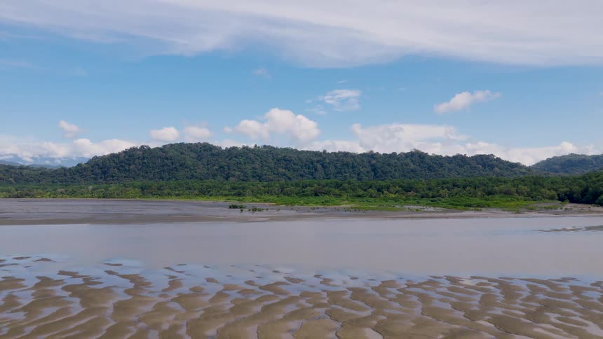 An aerial shot moves over a wide expanse of wet patterned sand and muddy water reflecting the bright blue sky revealing a dense forested mountain range in the distance