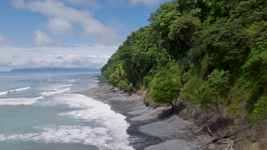 Waves roll in to shore and over the beach as trees and vegetation cover the promontory overlooking the coast