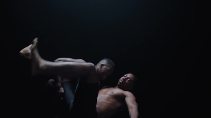 Two shirtless men engage in a grappling struggle on a blue mat under a dramatic spotlight in a dark room attempting to pin each other during a wrestling match