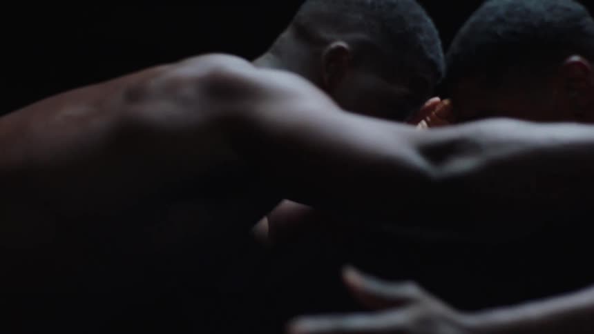 Two shirtless men grapple in slowmotion their bodies defined by stark light and shadow against a completely dark background