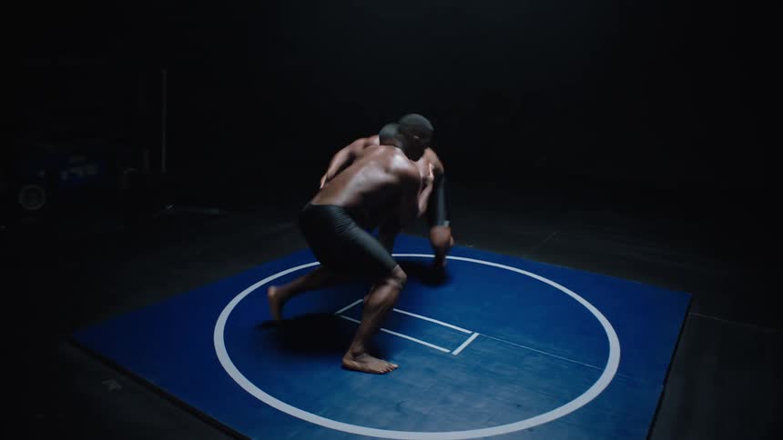 Two men wrestle on a blue mat in a dimly lit wooden floored room