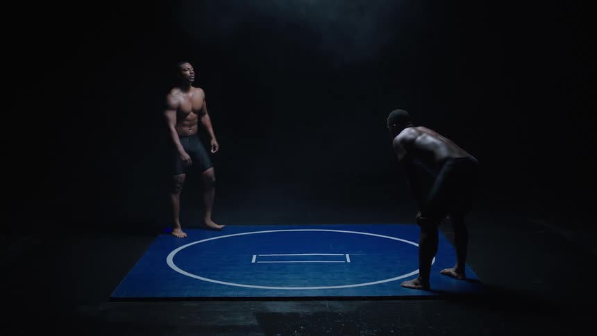 Two shirtless male wrestlers face each other in a crouched starting stance on a blue wrestling mat in a dimly lit setting poised for their match to begin