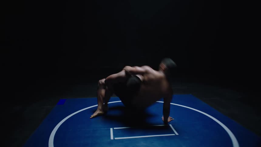 In a dimly lit studio two shirtless athletes grapple intensely on a circular blue mat spinning through the air before crashing down in a powerful takedown