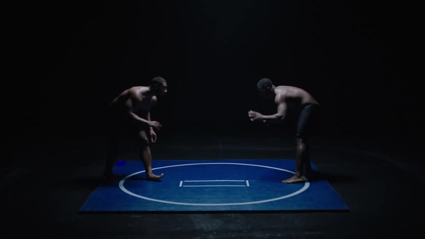 Two muscular men dressed only in shorts face each other on a dark blue wrestling mat in a dimly lit space they move into a wrestling stance circling each other briefly before one lunges forward to grapple