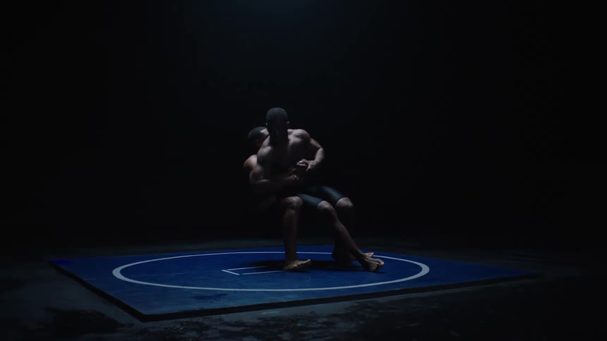Two athletes grapple on the ground on a blue mat under low lighting in a dark environment