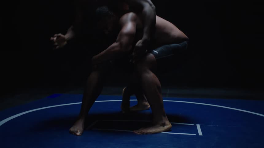 Two black men wrestle on a blue mate against a black background one wrestler takes down another in a throw to the mat