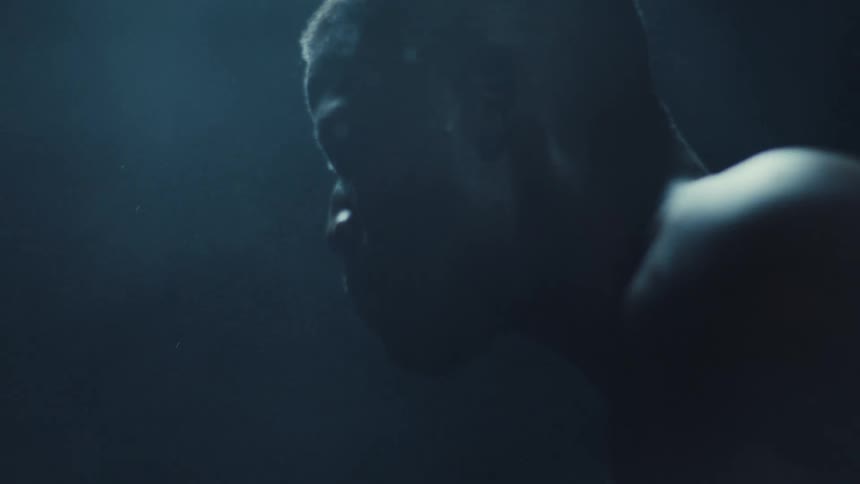 Bare chested man standing in a dimly lit room wipes his sweaty face