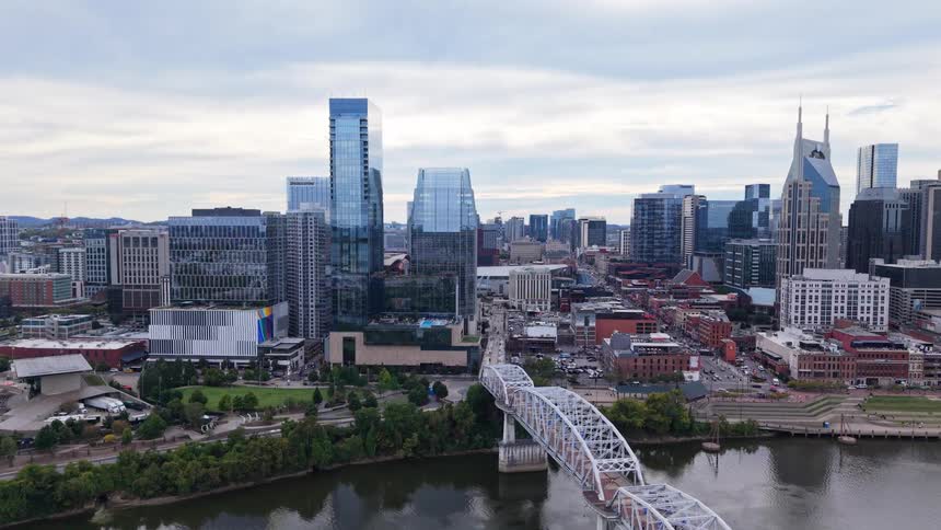 Gliding over a calm river and a white truss bridge the aerial shot moves toward a dense city skyline where towering glass skyscrapers stand out against a cloudy gray sky