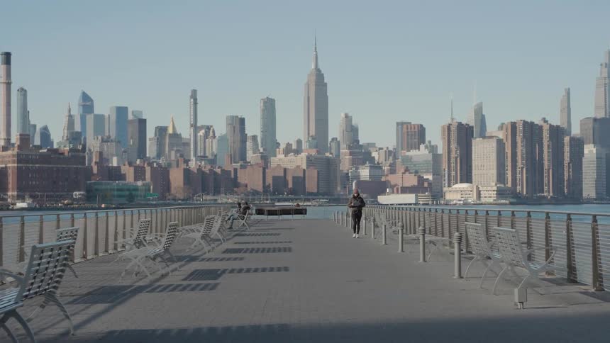 A woman walks along a wide empty pier with the iconic new york city skyline standing tall across the water under a clear bright sky