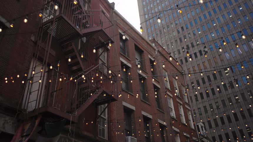 Urban alley with brick building and exterior staircase on the left hanging lights and modern building behind