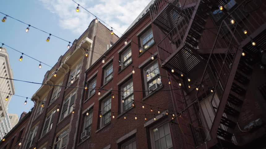 Old brick buildings with exterior staircase and hanging lights in urban alley under partly cloudy sky