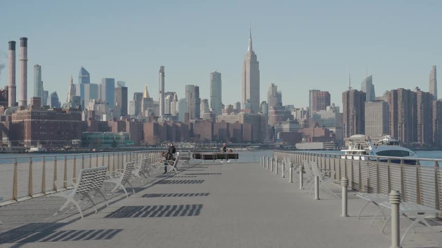 Several people jog and walk their bikes along a sunny pier as a ferry crosses the river with the vast new york city skyline filling the horizon