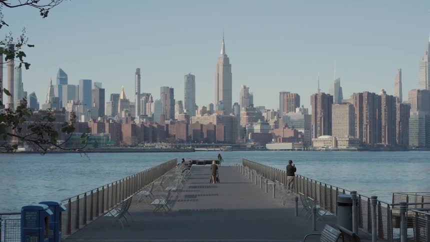 Several people walk along a long pier extending into the water with the iconic new york city skyline and the empire state building visible in the background