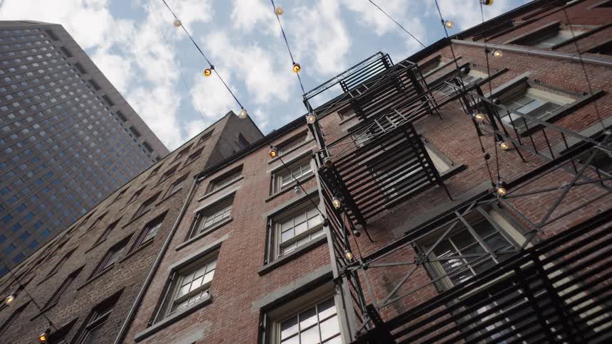 A low angle shot shows the brown brick exterior of an old apartment building with a black metal fire escape and string lights against a cloudy sky