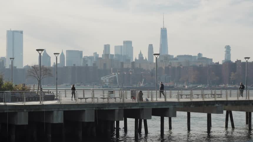 People stroll and walk their dogs along a waterfront pier on a hazy day with the sprawling city skyline visible in the distance across the water