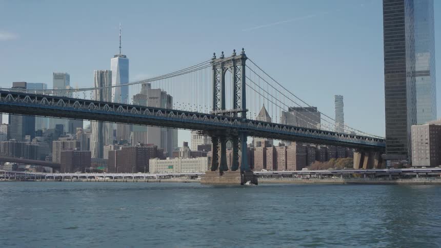 Cruising on the river the camera passes under a large bridge to reveal another bridge and the iconic downtown manhattan skyline on a bright clear day