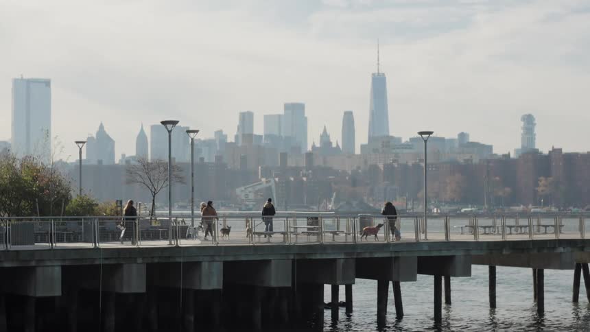 People walk their dogs and jog along a waterfront pier on a hazy day with the distant city skyline creating a picturesque backdrop