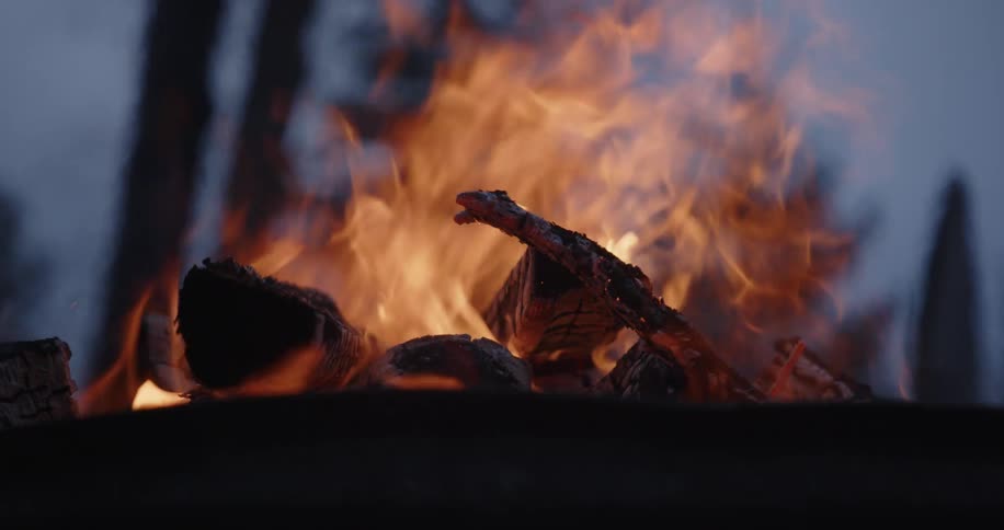 A low angle close view shows orange flames and smoke rising from burning wood chunks in a dark rusty metal fire pit outdoors