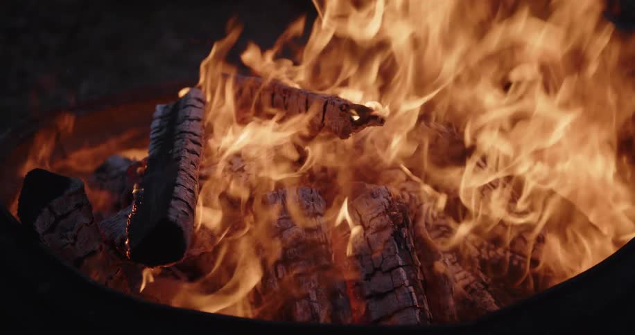 A close view of bright orange flames and smoke rising from a pile of burning wood logs in a dark metal fire pit