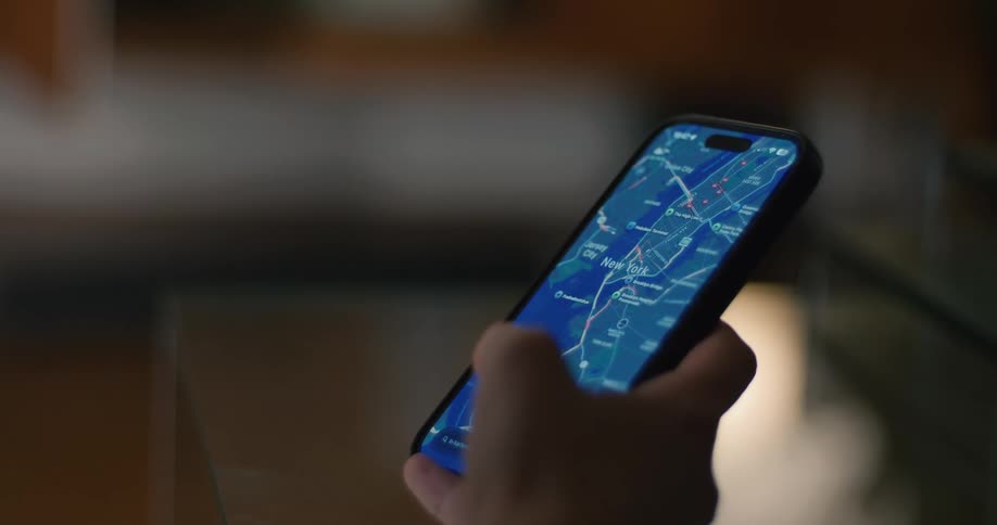 A person in a dim interior setting holds a smartphone looking at a digital map application centered on the new york city area displayed on the bright illuminated screen