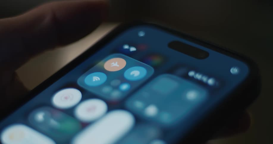 A persons finger taps the orange airplane mode icon on the control center screen of a glowing smartphone in a dark indoor environment