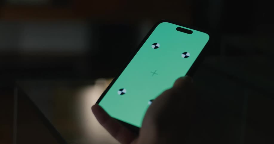 A persons hand holds a smartphone inside a dark room displaying a green screen