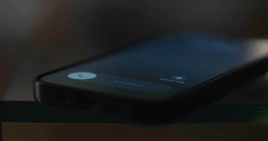 A smartphone lying faceup on a glass surface illuminates to display an incoming call screen vibrating slightly until a persons hand reaches in and picks up the device
