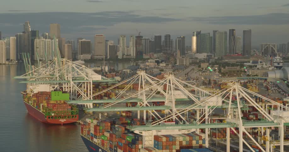 An aerial shot glides over a bustling shipping port where massive container ships are docked with the downtown city skyline visible across the water at sunrise