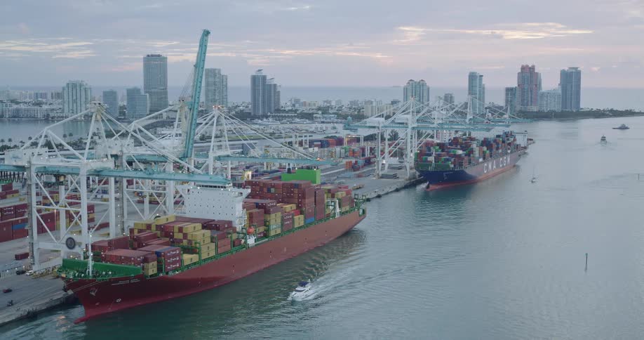 Aerial view of cargo ships docked at a busy container port next to a city skyline at dusk