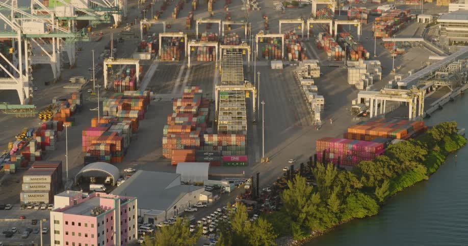 The video shows an aerial view of a large busy shipping port possibly in miami florida based on the skyline in the backgroundthe port is densely packed with stacks of colorful shipping containers and large container cranes situated right next to