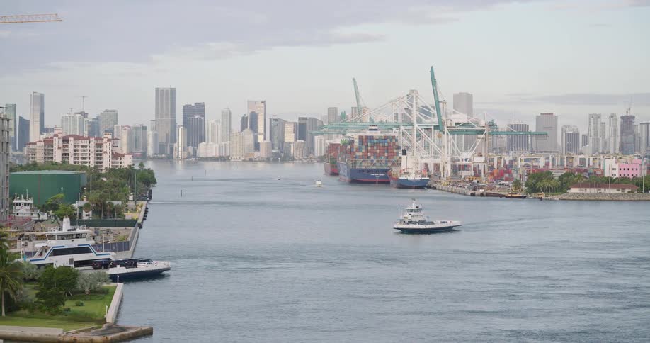 Urban channel connects a port area with cranes and cargo ships to a financial district with skyscrapers in the background