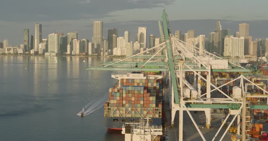 The video pans across a massive busy container port showcasing numerous towering gantry cranes and vast stacks of colorful shipping containersin the background the dense modern skyline of a major city is visible across the water with the port fac
