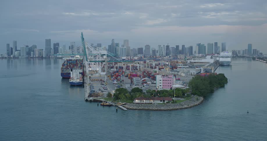 Port terminal with cranes containers and ships occupies an island surrounded by water with an urban skyline in the background