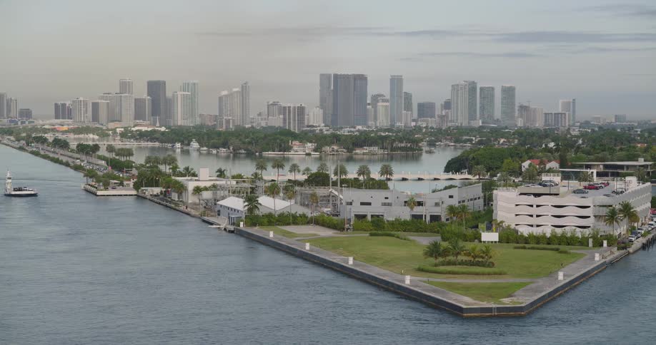 The aerial scene captures a prominent triangular spit of land surrounded by water featuring a lowrise white building and a parking structure leading the eye toward a raised bridge or causeway that connects to a dense background cityscape