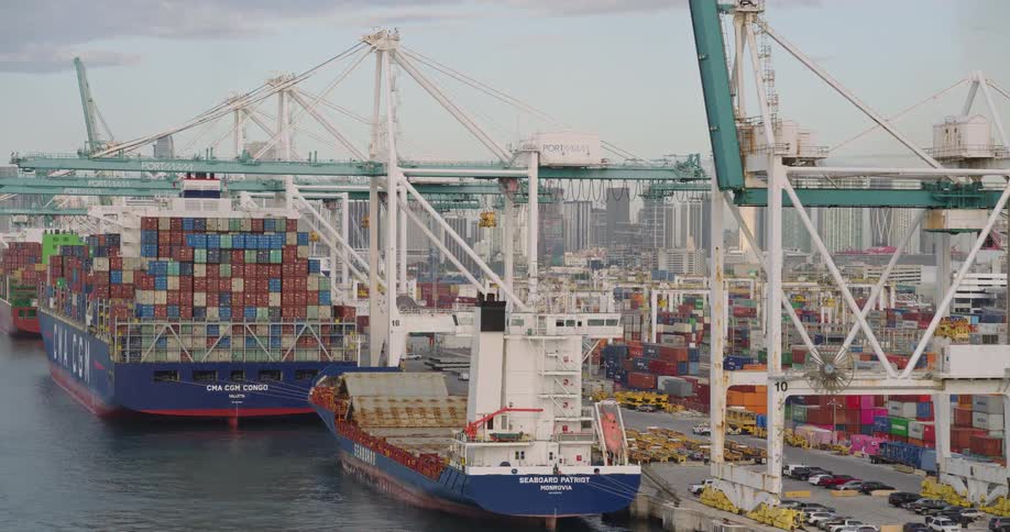 Flying alongside a bustling shipping port the aerial camera reveals massive cranes actively loading and unloading containers from docked cargo ships