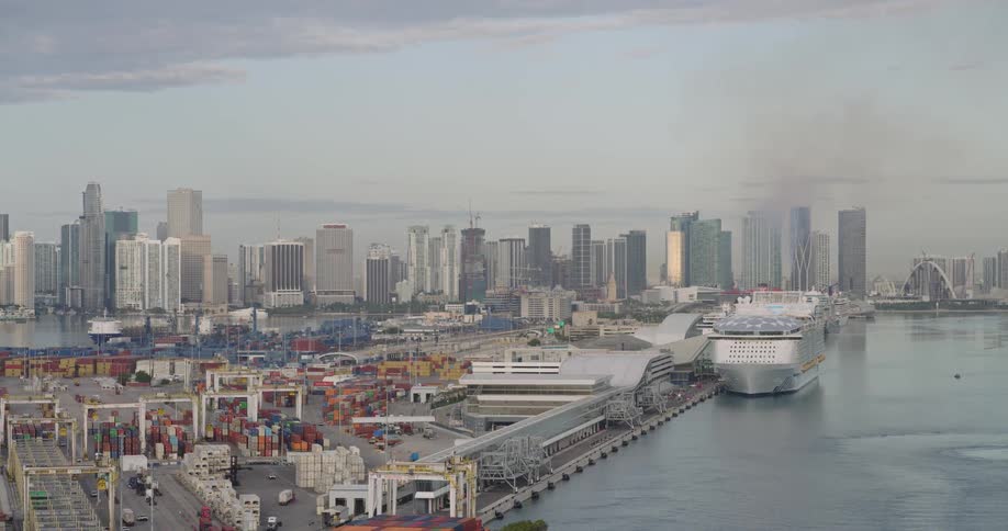The video shows a wide shot of a bustling port with a large cruise ship docked at a long pierin the background there is a dense skyline of modern skyscrapers suggesting a major metropolitan area like miami or a similar coastal city