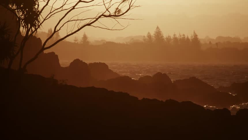 The video shows a warm sepiatoned silhouette of a rocky coastline at what appears to be sunrise or sunsetwaves crash against the dark rocks in the foreground with layers of trees and a hazy sky fading into the distance