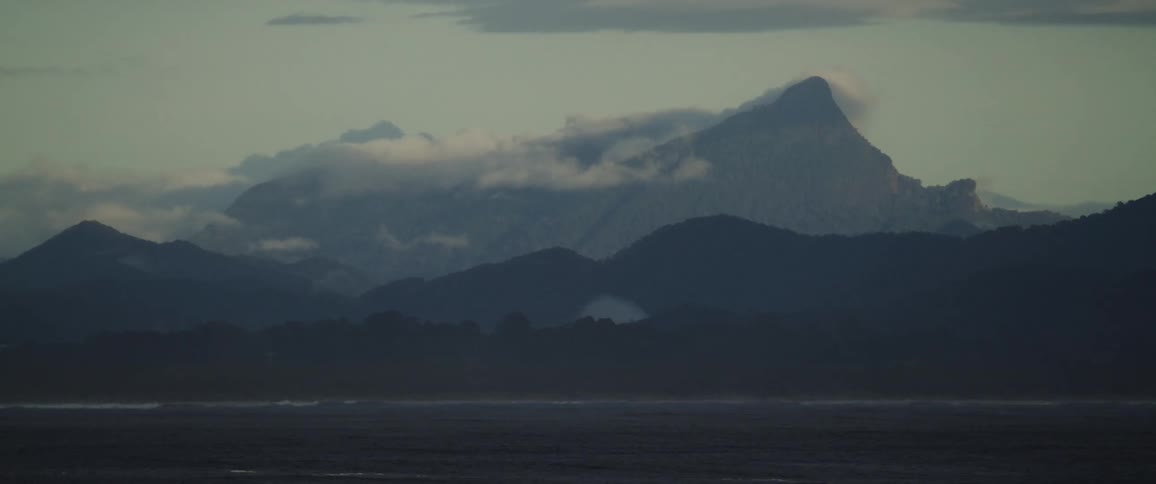 A rugged pointed mountain peak is silhouetted against a pale sky with clouds hanging over the forested slopes above a dark vast body of water