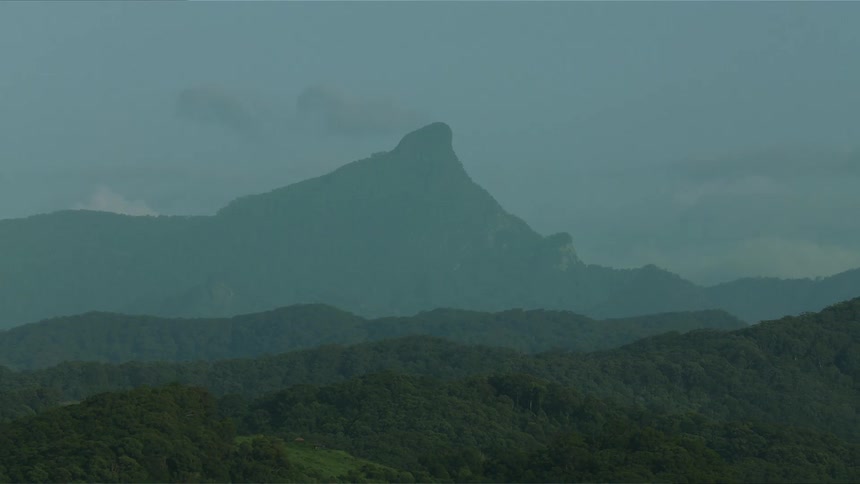 The video shows a static shot of a pointed mountain peak rising above densely forested hills the scene is slightly obscured by haze or mist giving it a cool desaturated bluegreen tone