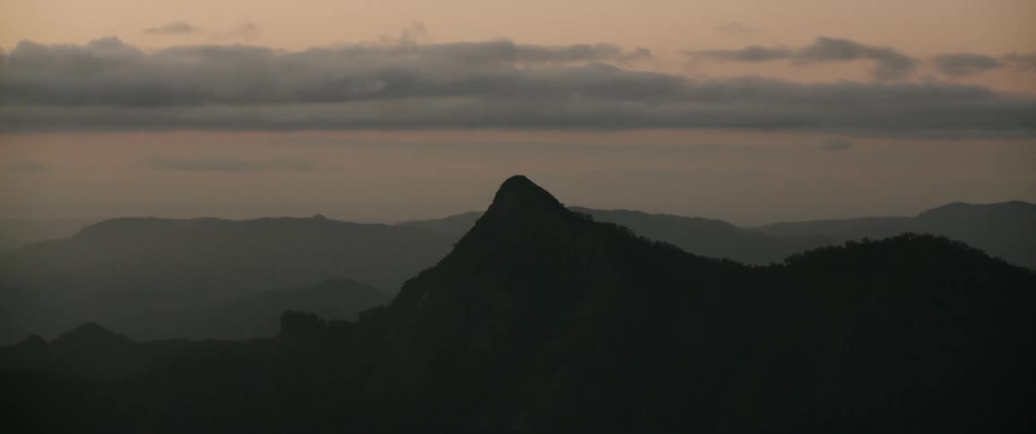 A dark pointed peak dominates the silhouette of distant mountains beneath a softly clouded dusky sky