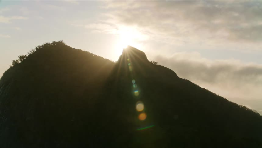 Mountain silhouette is outlined by the rising sun under a partly clouded sky