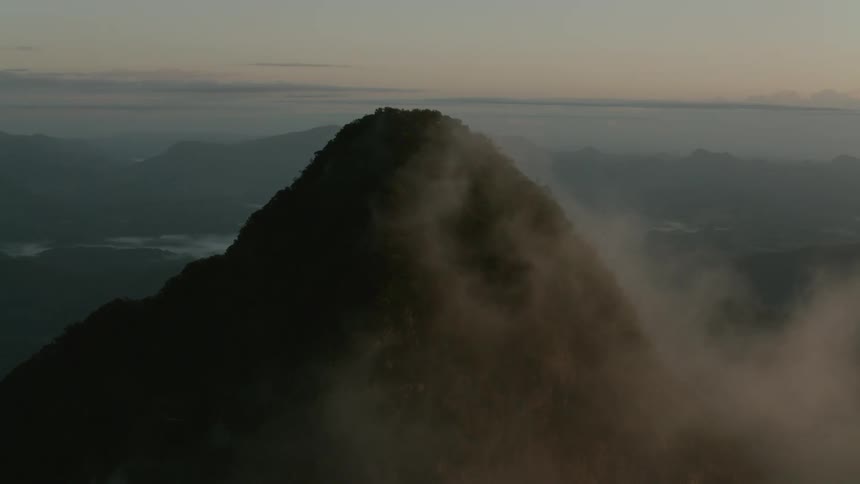 Peak of a mountain covered in fog with a valley and a mountain range on the background outdoors during dusk or dawn