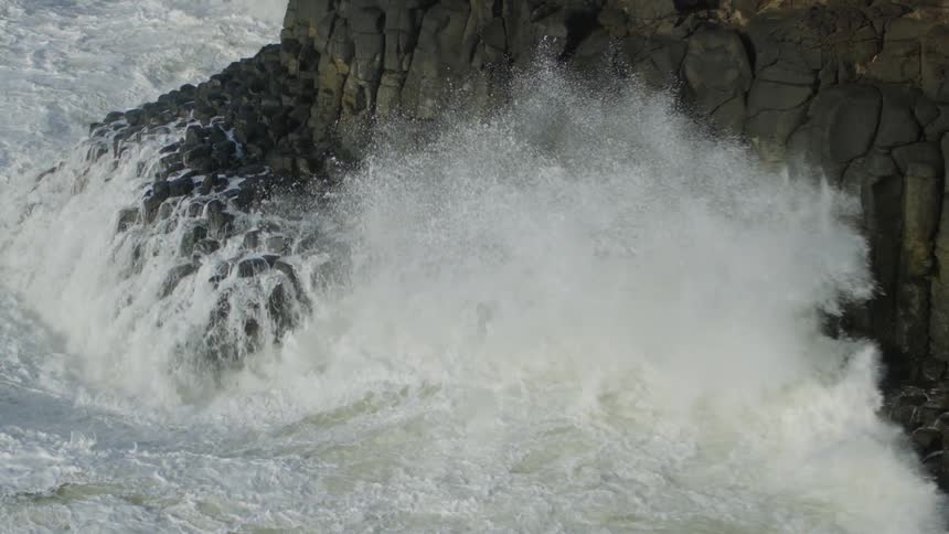 Powerful white waves crash against dark basalt columns on a rugged coastline