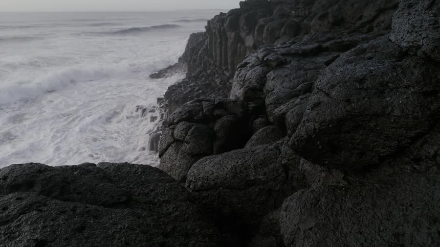 Powerful waves crash relentlessly against dark jagged volcanic cliffs under an overcast sky