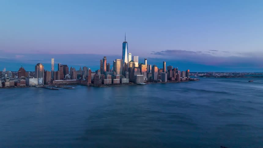 An aerial wideangle establishing shot of a city skyline in a time lapse scene featuring prominent skyscrapers and reflecting the blue and pink light over a wide river surface
