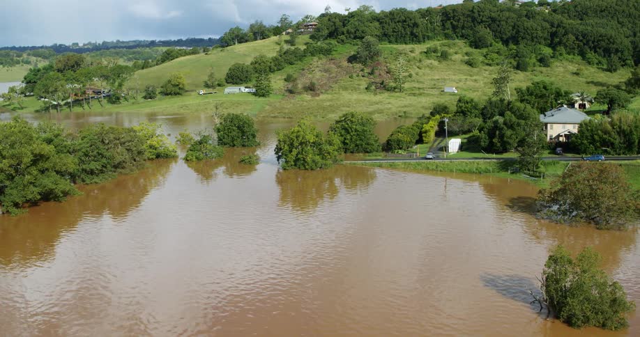 This aerial shot captures a valley landscape where extremely high floodwaters murky brown in color have submerged the lower areas a rural road running along the edge of the flood is visible with a small number of cars driving past houses nestled on th