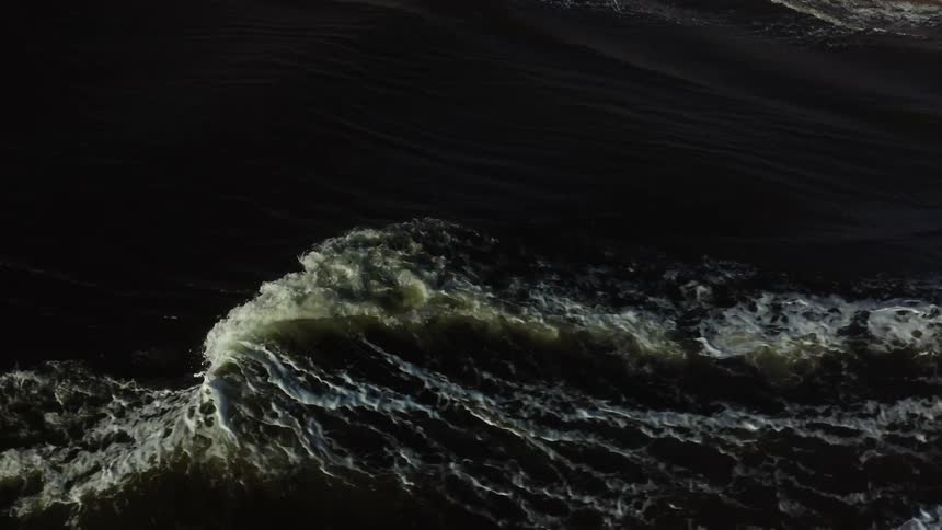 An intense highangle aerial view of a large dark ocean wave cresting and breaking with the bright white foam contrasting sharply against the surrounding deep tumultuous water