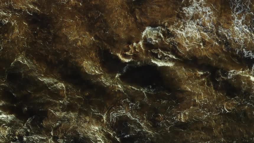 An aerial view captures the relentless motion of murky brown water as it swirls and surges with whitecaps breaking on the surface