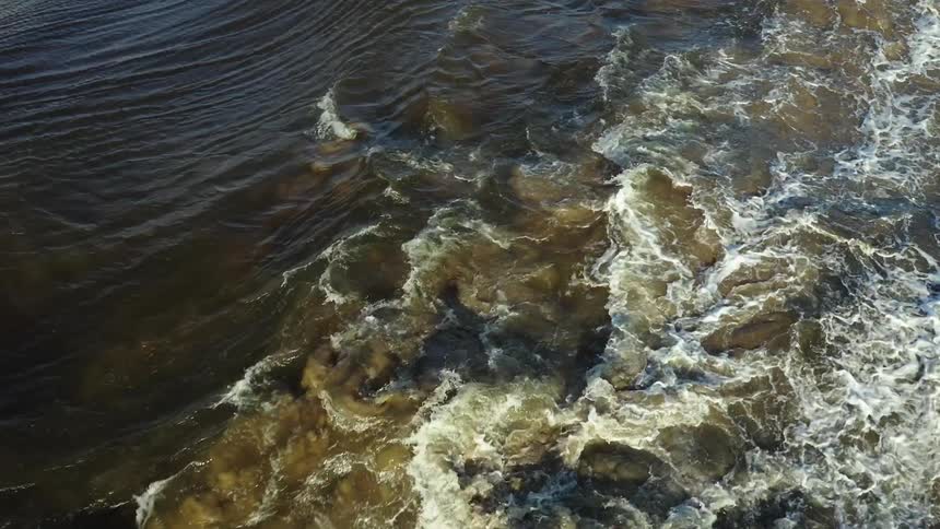 Flowing water forms whirl patterns and white foam on the surface of a river seen from above