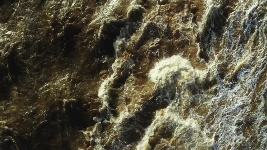 An aerial closeup view captures the turbulent dark sedimentladen water of a river heavily churning and producing white foam